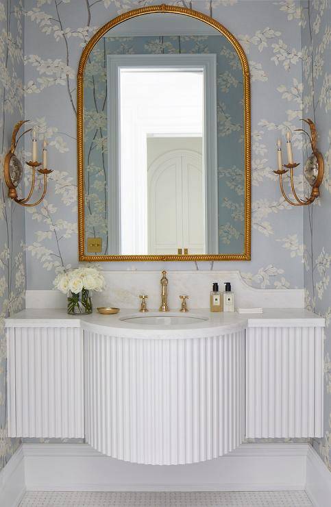 gold arch mirror in half bathroom with white pole wrap sink gold candle wall sconces with floral wallpaper