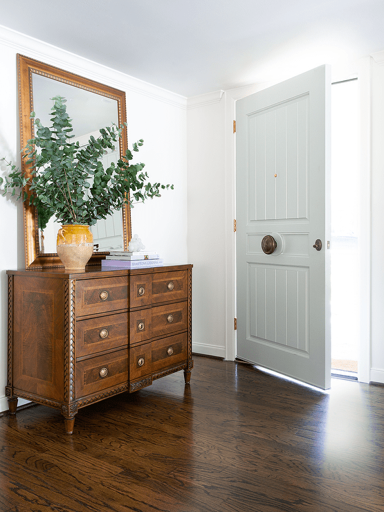 A gold beveled mirror sits atop an antique French chest placed in an entryway near a white front door.