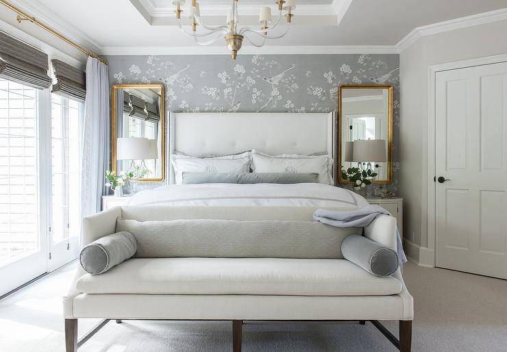 Gold leaf mirrors over white French nightstands in a tony white and gray bedroom finished with crystal lamps. A white settee with gray perpetuate pillows is finished at the foot of the bed with a sophisticated touch.