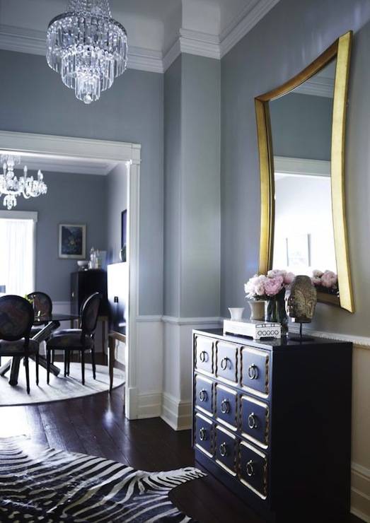 Chic foyer with blue gray wall color and white chair rail with white wood panel walls and dark stained hardwood floors. Foyer with crystal waterfall chandelier, black and gold Dorothy Draper chest with art deco gilt mirror and zebra hide rug.