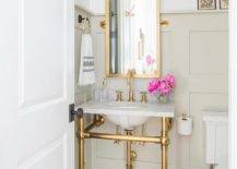 A marble and brass washstand is fixed against a light taupe wainscot wall to green and black cement mosaic floor tiles. Above the sink vanity, a tall brass pivot mirror is lit by a brass and gold 2-light sconce.