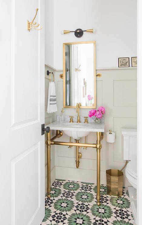 A marble and brass washstand is fixed against a light taupe wainscot wall to green and black cement mosaic floor tiles. Above the sink vanity, a tall brass pivot mirror is lit by a brass and gold 2-light sconce.