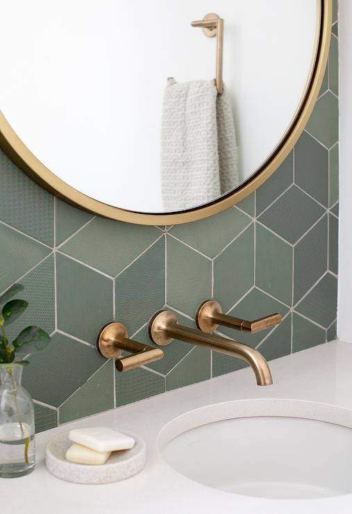 round gold mirror hangs from a wall covered in green geometric tiles over a brushed gold faucet fixed above a round sink.