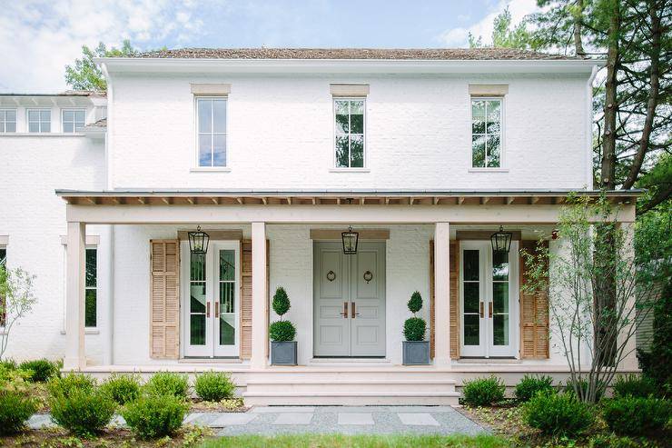 Stunning white brick cottage home with soft gray French front doors, vintage wood shutters and a covered porch with decorative plants.