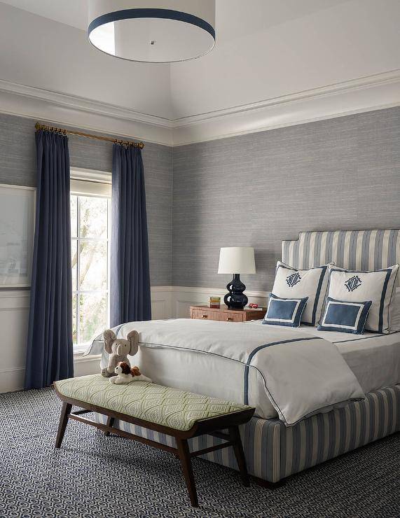 gray and blue bedroom features a dark stained wood bench with a green diamond print cushion placed on a blue and gray carpet at the foot of a gray striped bed topped with white and blue hotel bedding accented with blue monogrammed pillows. The bed sits against a gray grasscloth wallpapered wall lined with white wainscoting. The room is finished with a navy blue double gourd lamps placed on brown nightstands and a window covered in blue curtains.