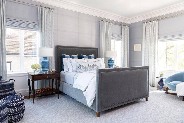 gray and blue bedroom charcoal gray velvet bed placed against a wall clad in gray check wallpaper and positioned between cherry French nightstands topped with blue stripe lamps. The bed, accented with blue plaid pillows and blue hotel bedding, sits on a gray rug, while windows are covered in gray roman shades layered behind gray curtains.