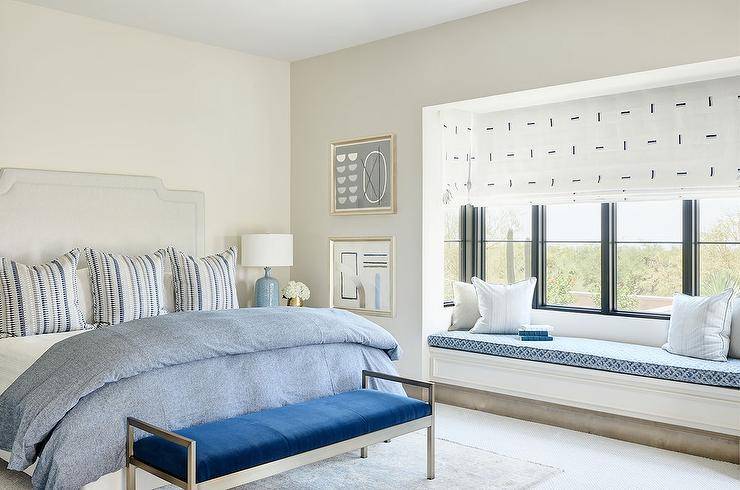 Light gray and blue bedroom features a light gray French headboard accented with gray and blue stripe pillows, a blue bench at the foot of the bed and a bedroom nook white built in window seat with blue cushion.
