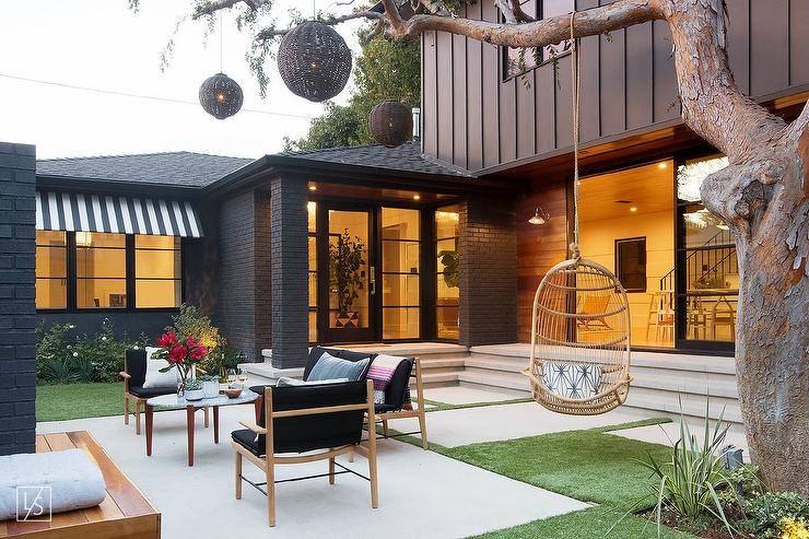 contemporary home sunken patio featuring a mature tree with a hanging woven chair and black woven chandeliers. Black outdoor chairs and sofa frame an oval teak and marble coffee table with colorful floral arrangements.