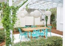 A white pergola holds staggered white chandeliers over a teak outdoor dining table paired with aqua blue patio dining chairs.