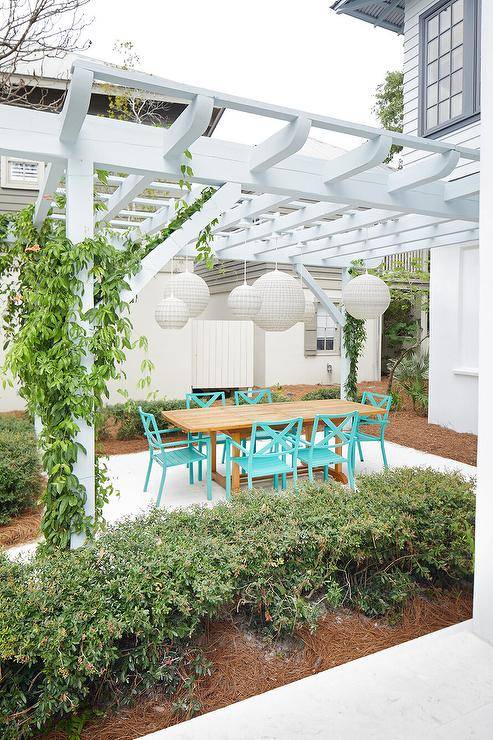 A white pergola holds staggered white chandeliers over a teak outdoor dining table paired with aqua blue patio dining chairs.