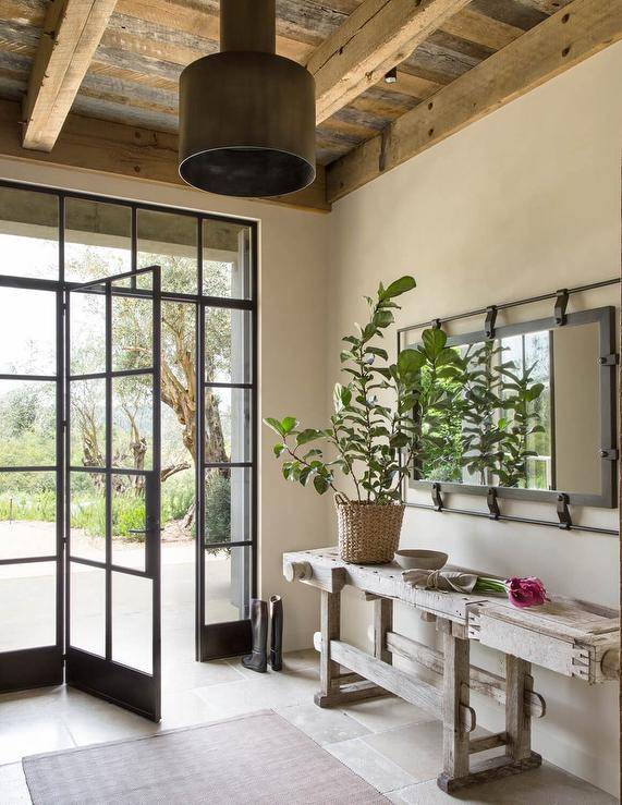 Cottage style foyer features a reclaimed wood console table under a long rectangular iron mirror, a steel and glass front door and a plank ceiling with wooden beams.