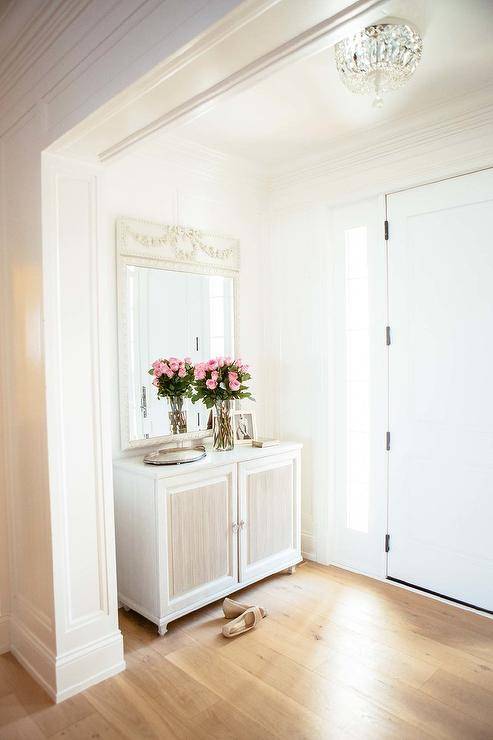 An ivory French mirror in a white, well-lit foyer paired with a white cabinet with reeded doors and glass knobs. A crystal flush mount light delicately decorates the entryway over warm wood oak plank flooring.