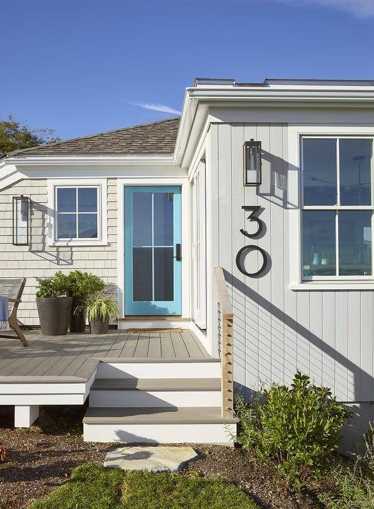 Cottage home features a turquoise blue front door with glass panels and gray siding completed with modern black house numbers. A gray teak deck keeps a neutral surface with wood and wire railing.