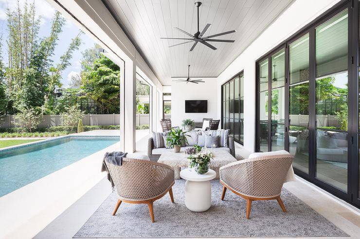 overed patio with 2 sitting areas that boast woven chairs and a round white accent table, and black fans mounted on a gray plank ceiling, next to an in-ground pool.overed patio with 2 sitting areas that boast woven chairs and a round white accent table, and black fans mounted on a gray plank ceiling, next to an in-ground pool.