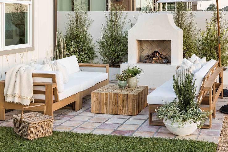 Cottage patio design features beige teak outdoor sofas with white cushions, a reclaimed wood coffee table, a stucco outdoor fireplace and a wicker picnic basket.