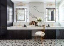Chic gray and black bathroom boasts a gold metal French chair placed on gray and black hexagon floor tiles at a black dual donning black pulls and a thick marble countertop. Aged brass faucets are fixed in front of a marble backsplash and beneath vanity mirrors mounted in front of windows.