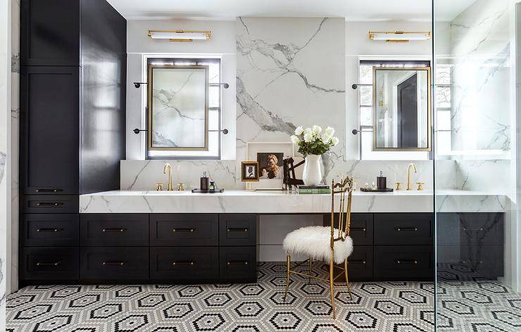 Chic gray and black bathroom boasts a gold metal French chair placed on gray and black hexagon floor tiles at a black dual donning black pulls and a thick marble countertop. Aged brass faucets are fixed in front of a marble backsplash and beneath vanity mirrors mounted in front of windows.
