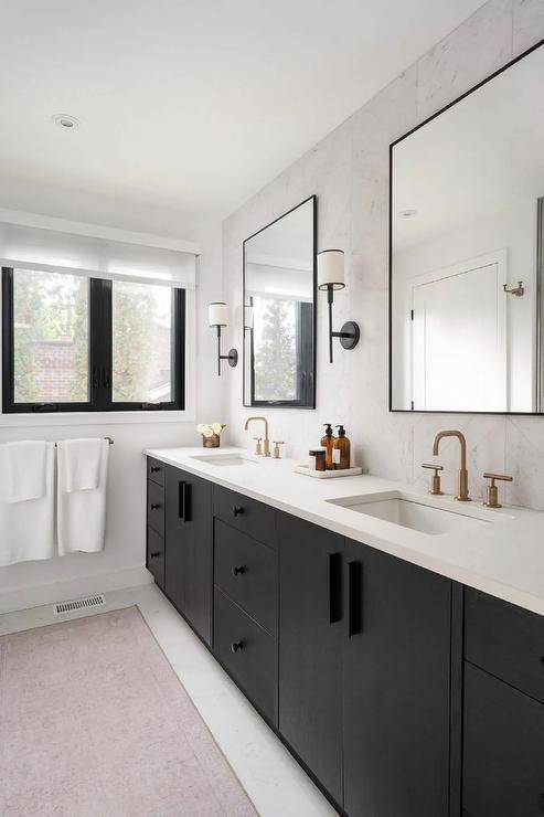 A pink runner sits in front of a black dual bath vanity accented with black hardware and brushed gold faucets fixed under black framed mirrors lit by oil rubbed bronze sconces.