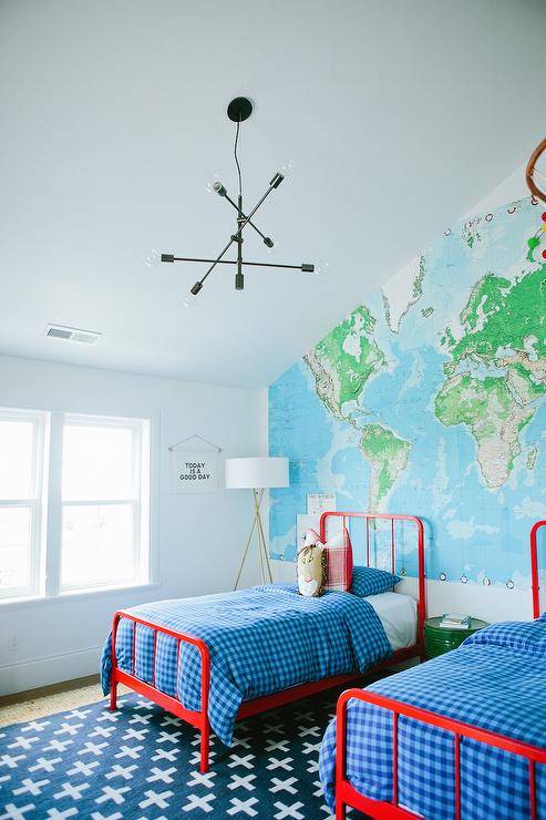 Red and blue boys' bedroom features a sloped ceiling over an accent wall lined with a World Map Paper Mural lined with red twin beds, Land of Nod Primary Bed, dressed in blue buffalo check bedding and red buffalo check pillows flanking a round green metal drum table doubling as a nightstand atop a Land of Nod Indoor + Outdoor Rug.