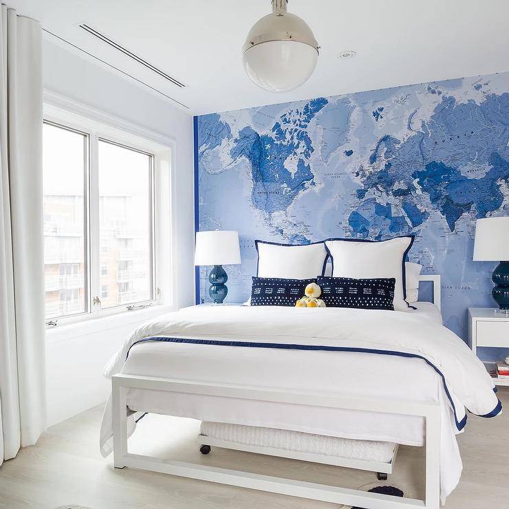 Blue and white boy's bedroom features a white Parsons bed with a navy blue border duvet and navy blue border shams on blue world map wallpaper, blue double gourd lamps on white nightstands, illuminated by a nickel and white Hicks Pendant.