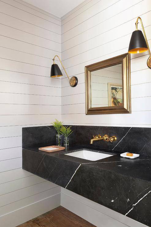 Fixed to shiplap trim, gold and black sconces flank a square gold beveled mirror is hung over a honed black marble floating sink vanity finished with a brass faucet kit fixed to a black marble backsplash.
