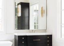 Marble and brass sconces on a frameless beveled vanity mirror above a black washstand with a white quartz countertop and brass pulls.