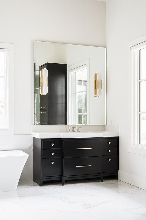 Marble and brass sconces on a frameless beveled vanity mirror above a black washstand with a white quartz countertop and brass pulls.