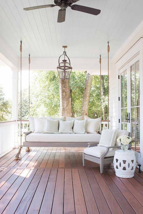 Gorgeous cottage porch features a gray wash porch swing sofa hanging by ropes from a white beadboard ceiling and topped with a light gray cushion and white border gray white border pillows accented by a matching accent chair sat beside a white rope stool lit by a antiqued brass lantern hanging over a composite porch board floor.