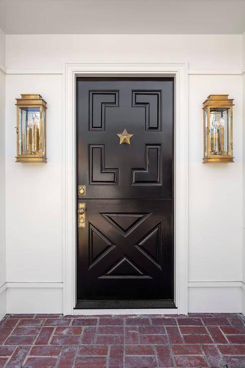 Brass carriage sconces flank a black Dutch geometric door at a front door with red brick pavers on porch surface.