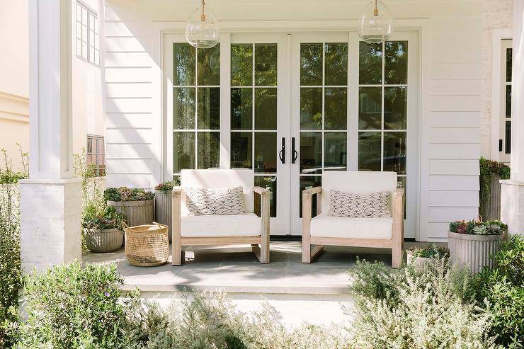 styled covered porch boasts hand blown glass lanterns hung over Restoration Hardware Aegean Teak Chairs placed beside gray planters and in front of sliding glass doors framed by white siding
