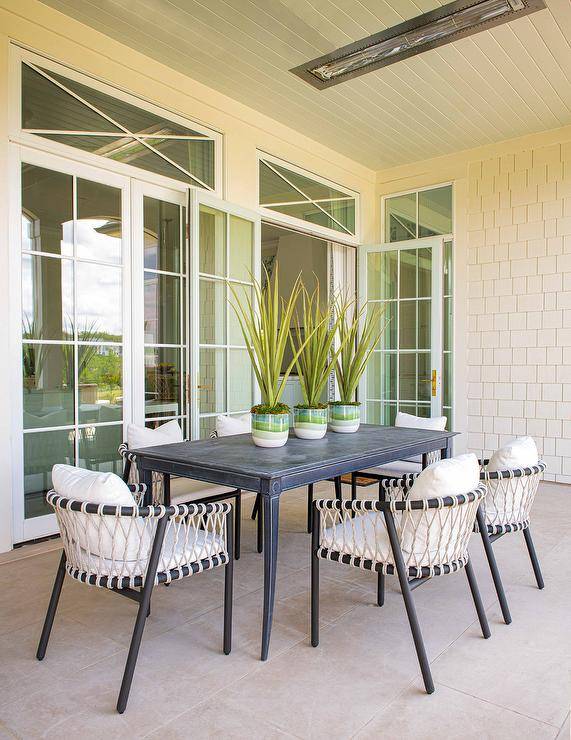 Black and white rope chairs sit at a black French outdoor dining table placed on a covered patio and warmed by a ceiling-mounted heater.
