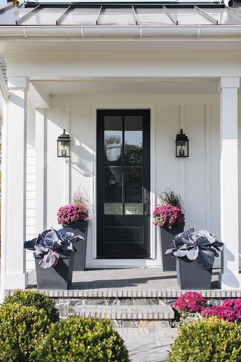 Modern black planters sit on a covered porch complementing a white home exterior contrasted with a glass paneled black front door flanked by iron carriage lanterns.