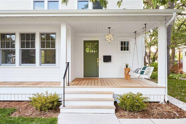 An olive green front door accents a white home fitted with white siding and a Moravian star pendant hung from a white plank covered porch ceiling in front of a white rope swing.