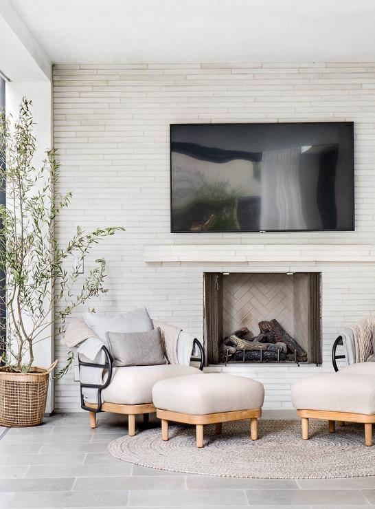 Spacious covered patio features white fireplace wall split face tiles under a mounted TV and an off white accent chair with off white ottoman atop a tan rug.