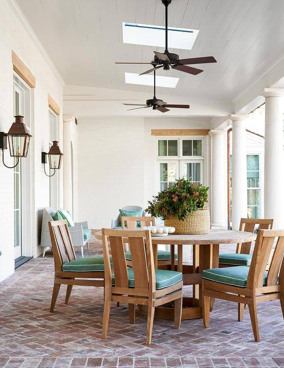 Ceiling fans mounted to a white plank ceiling with skylights cool a covered patio boasting a round teak dining table surrounded by brown teak chairs placed on red brick herringbone pavers.