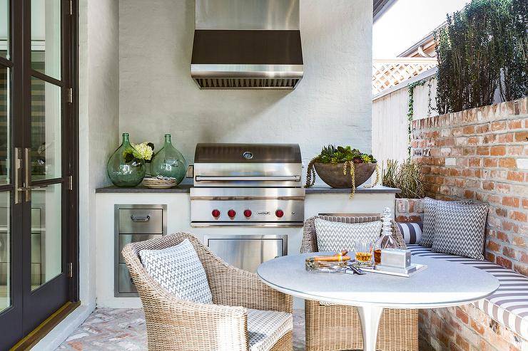 Outdoor patio designed with a red brick bench and wall upholstered with a gray and white striped cushion. A round outdoor dining table is paired with a set of wicker chairs next to a cooktop designed with a small wolf bbq, stainless steel vent hood, and kitchen space.