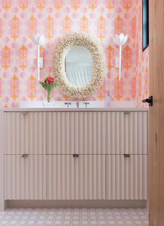 contemporary bathroom features a blush pink washstand fitted with a polished nickel faucet kit mounted under an oval coral mirror hung between white flower sconces from a wall covered in pink and orange wallpaper.