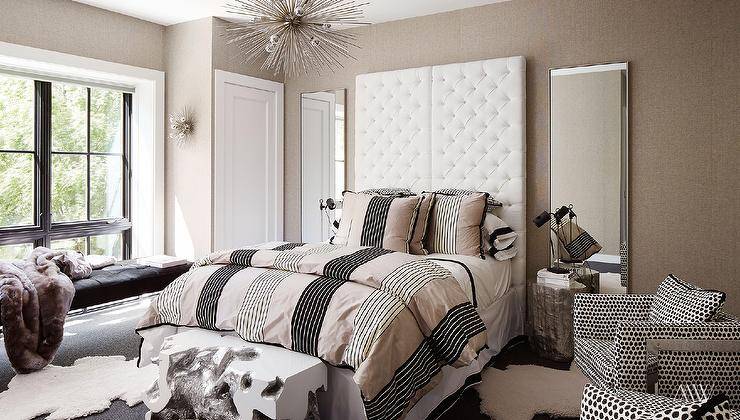 Full length mirrors are mounted against a light taupe wallpapered wall behind silver faux bois bedside tables and flanking side-by-side all white tufted headboards. The headboards support a bed dressed in beige and black bedding lit by a silver sputnik chandelier. A black and white accent chairs sits on a white sheepskin rug beside the bed.