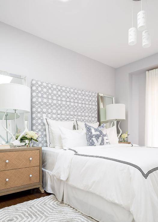 Stunning bedroom features a light gray wall lined with a black and white geometric shower curtain stapled onto a headboard on bed dressed in black and white hotel bedding flanked by woven nightstands topped with white antler lamps and beveled mirrors.