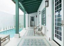 A gorgeous long green plank covered patio is accented with green and aqua blue mosaic floor tiles fitted under light gray teak chairs topped with white and gray pillows. The chairs are placed beneath a white framed windows with a green trim and positioned facing a lap pool.