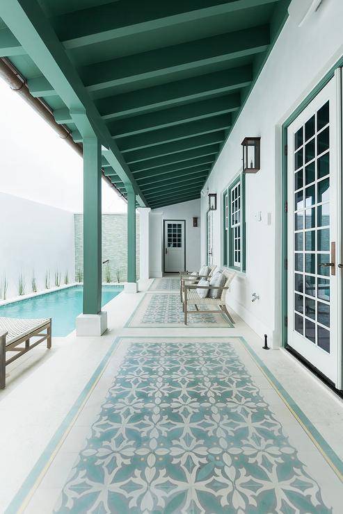 A gorgeous long green plank covered patio is accented with green and aqua blue mosaic floor tiles fitted under light gray teak chairs topped with white and gray pillows. The chairs are placed beneath a white framed windows with a green trim and positioned facing a lap pool.