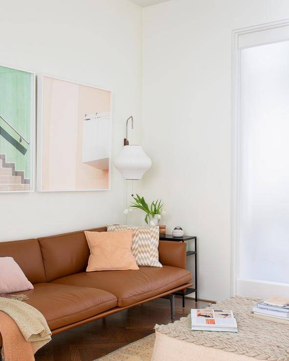 Transitional living space furnished with a brown leather sofa accented with various pastel pillows and a pastel-colored wall art to complement the tones. Wood herringbone pattern floors invite an updated and charming appeal finished with a rug and woven pouf as a coffee table.