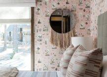 A pink fringe mirror hangs from a pink wallpapered wall beside a window dressed in a white roman shade and facing a gray upholstered bed complemented with pink and gray plaid bedding.