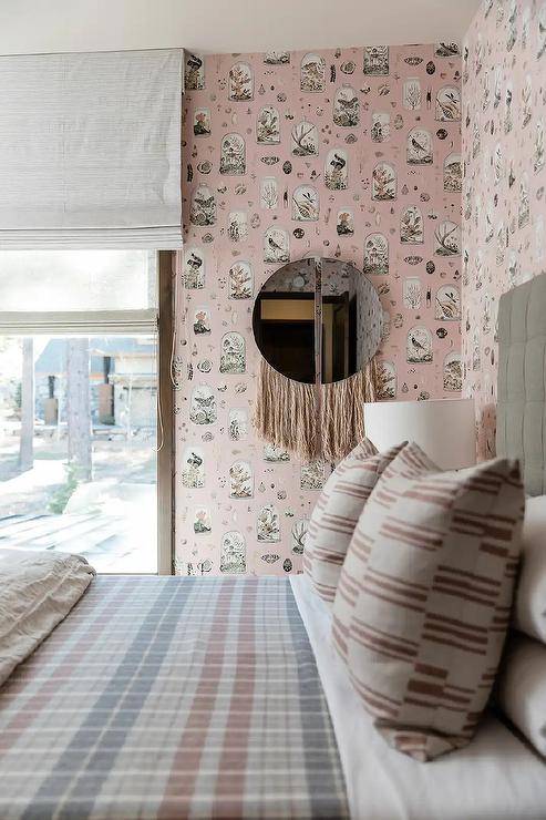 A pink fringe mirror hangs from a pink wallpapered wall beside a window dressed in a white roman shade and facing a gray upholstered bed complemented with pink and gray plaid bedding.