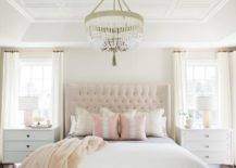 white and pink bedroom boasts white greek key nightstands topped with pink glass lamps and placed beneath windows dressed in white curtains hung from brass rods beneath a tray ceiling accented with white decorative trim. The nightstands flank a pink tufted wingback headboard complementing a bed covered in white bedding paired with a pink faux fur throw and pink pillows