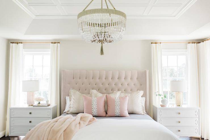 white and pink bedroom boasts white greek key nightstands topped with pink glass lamps and placed beneath windows dressed in white curtains hung from brass rods beneath a tray ceiling accented with white decorative trim. The nightstands flank a pink tufted wingback headboard complementing a bed covered in white bedding paired with a pink faux fur throw and pink pillows