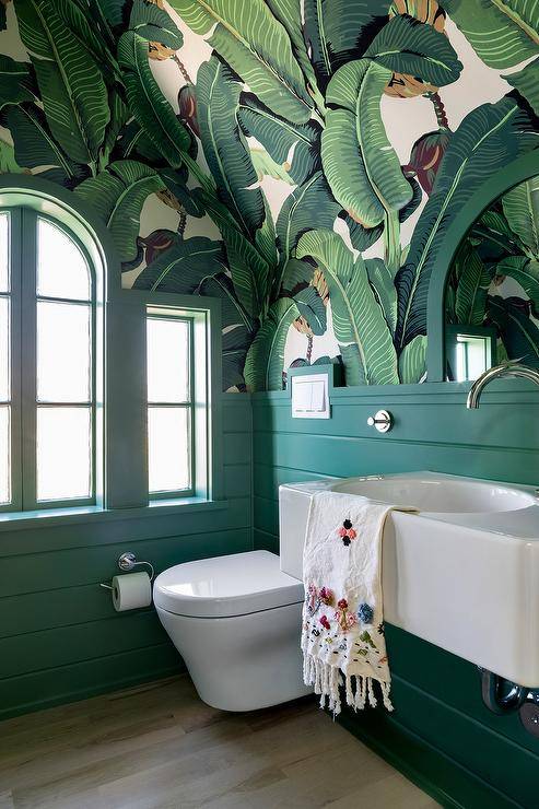 A white floating sink is fixed to a green shiplap backsplash beside a floating toilet in a goregous contemporary powder room. Upper walls are clad in palm leaf print wallpaper that continues up to a vaulted ceiling.