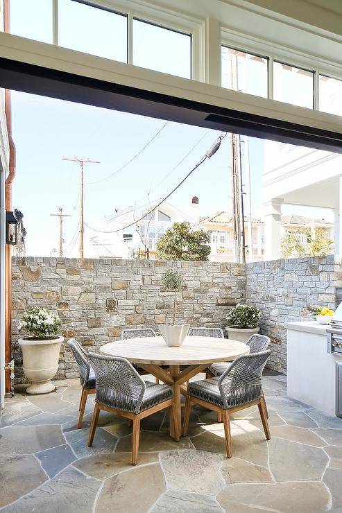 Candelabra Home Loom Dining Chairs sit around a round wooden dining table placed on gray pavers framed with a gray stone privacy wall.