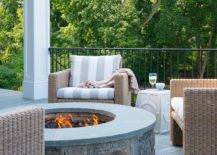 Cozy round fire pit, made of concrete and slate tiles is surrounded by outdoor wicker chairs with white and gray striped cushions. Iron patio railing closes off the space overlooking lush green views.