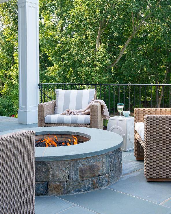 Cozy round fire pit, made of concrete and slate tiles is surrounded by outdoor wicker chairs with white and gray striped cushions. Iron patio railing closes off the space overlooking lush green views.